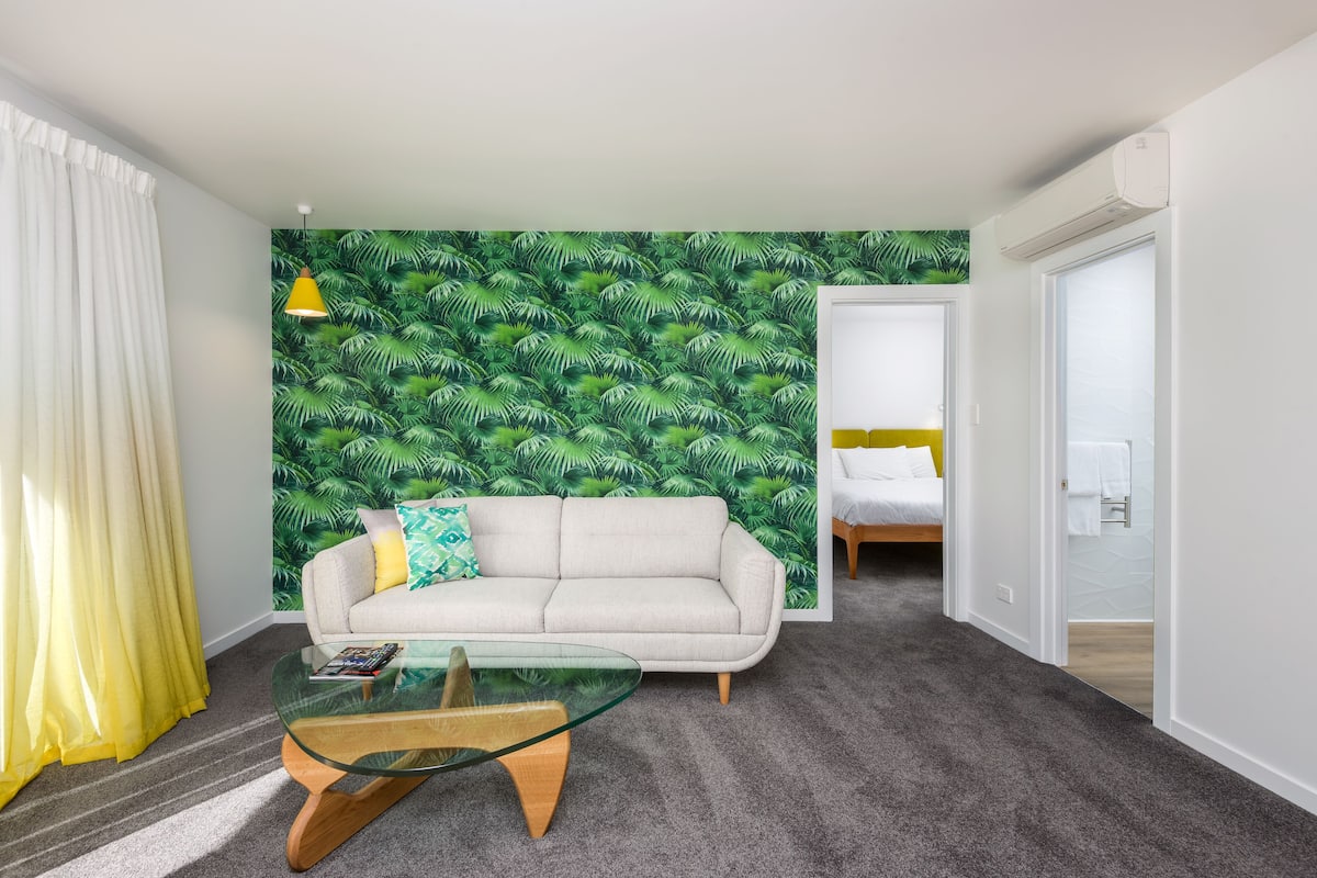 A modern room with a green leaf-patterned wall, a sofa, a glass coffee table, and a bed in the background.