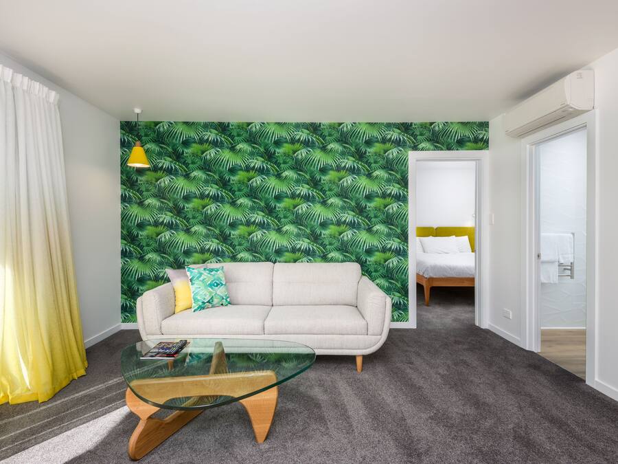 A modern room with a green leaf-patterned wall, a sofa, a glass coffee table, and a bed in the background.