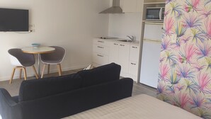 A compact room with a black sofa, a small round table with two chairs, a kitchenette with a microwave, and a TV mounted on the wall.