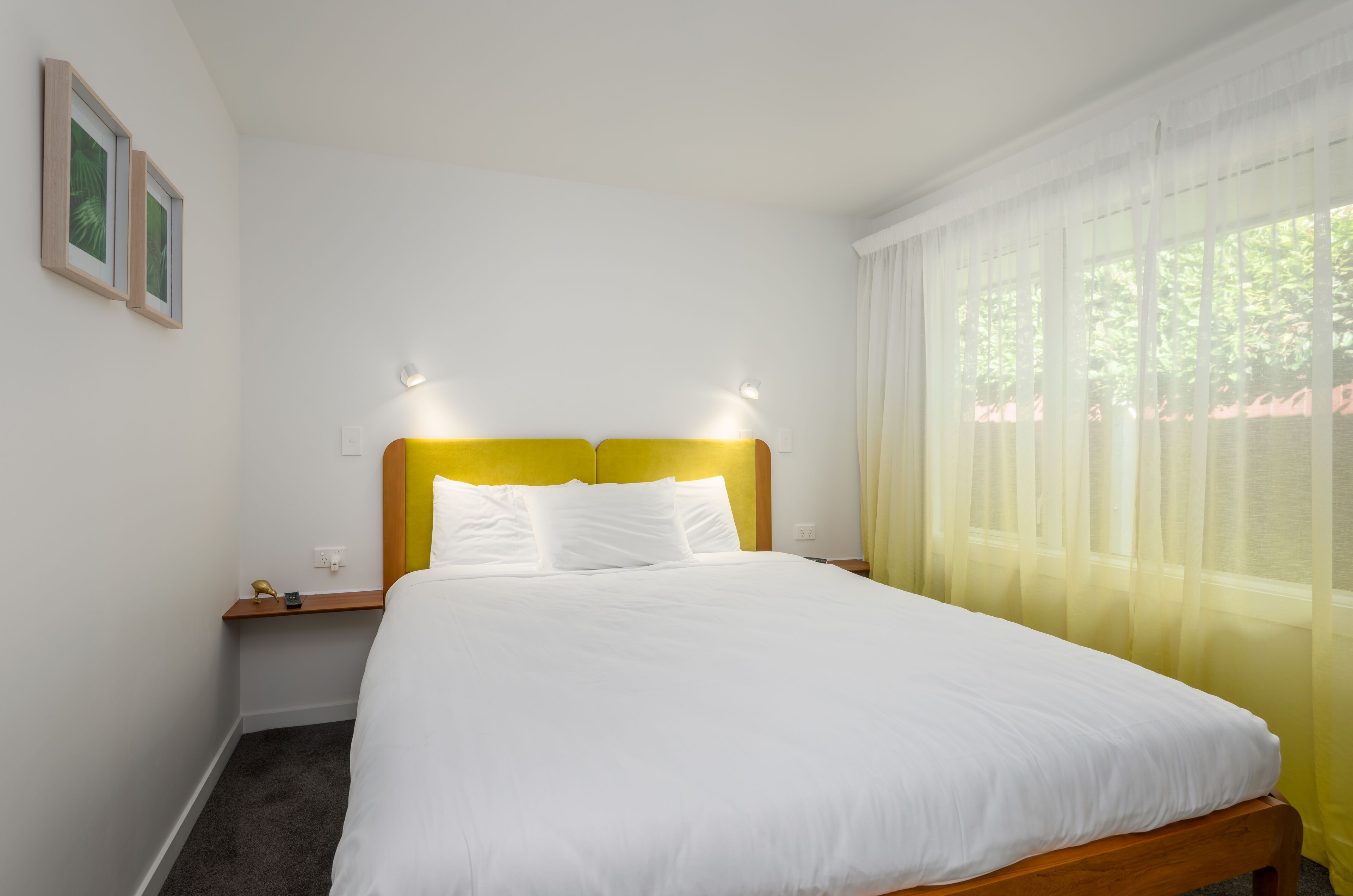 Una cama bien tendida con ropa de cama blanca y un cabecero amarillo, una mesita de noche con una lámpara y una ventana con cortinas translúcidas.