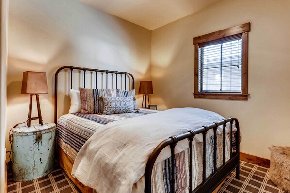 A bedroom with a bed, two bedside lamps, a window with blinds, and a small blue container. - 2 Blocks To Grand Lake: Downtown Western Cabin! (Grand Lake)