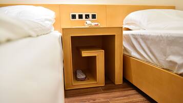 Habitación de hotel con dos camas, una mesita de noche de madera con un teléfono y un enchufe, y un pequeño espacio de almacenamiento.