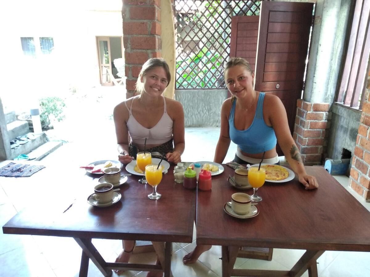 Dos personas sentadas en una mesa de madera con platos de desayuno, vasos de zumo de naranja y un muro de ladrillo al fondo.