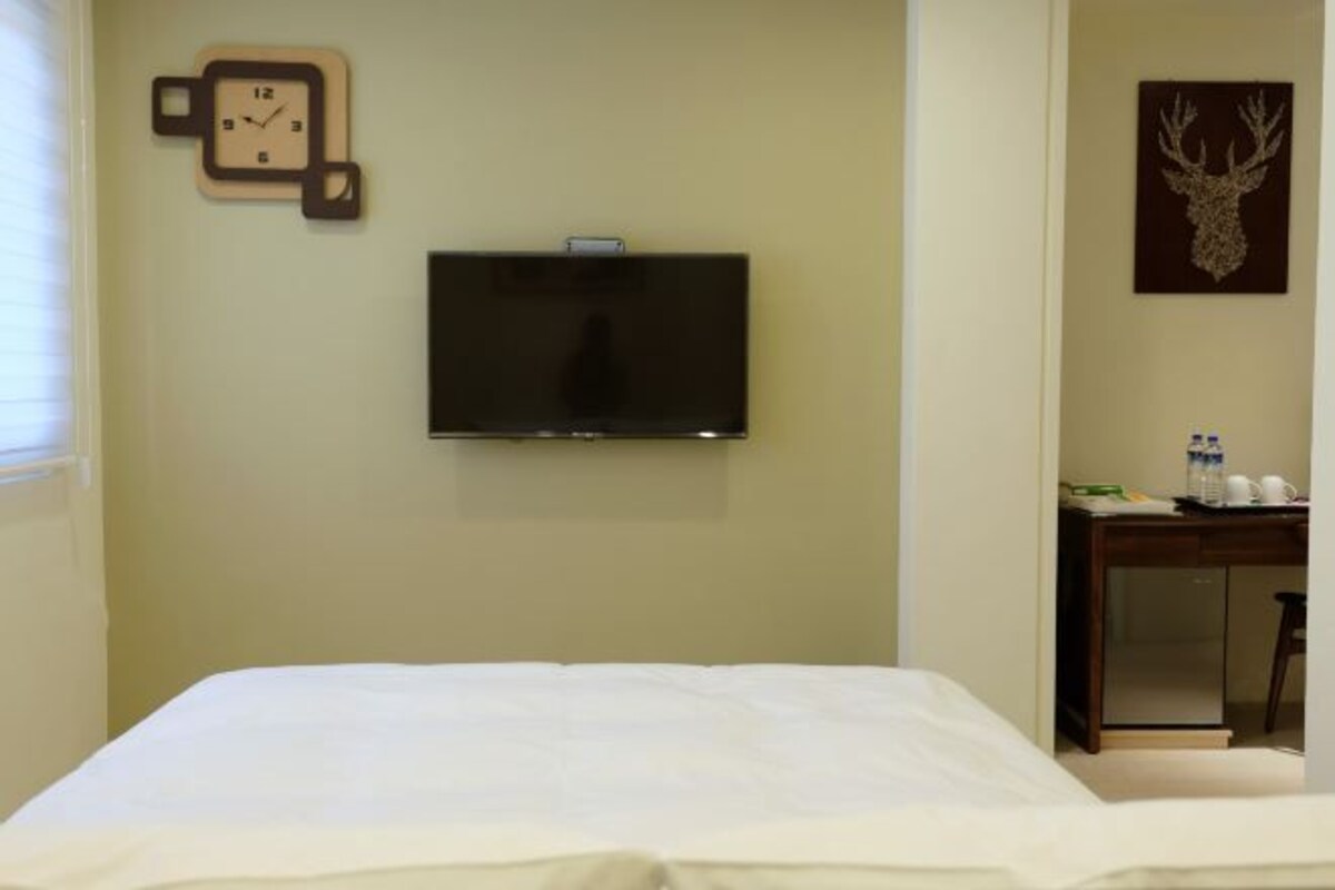 A hotel room with a bed, a television mounted on the wall, a clock, and a deer head decoration.