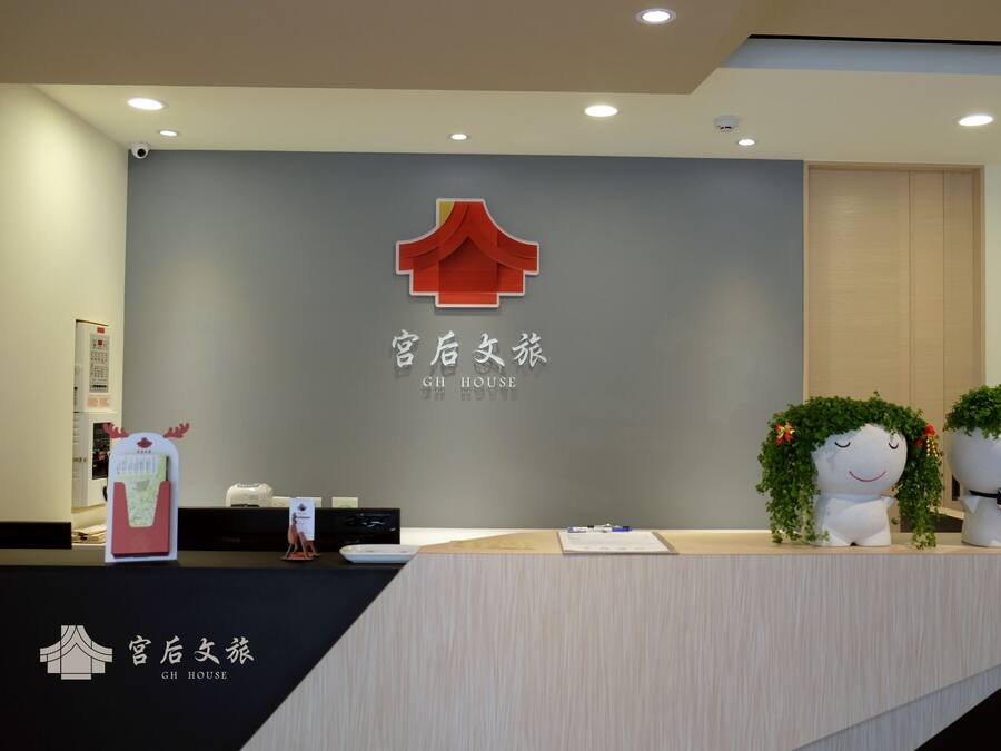 A hotel reception desk with a logo, a wall-mounted sign, and two decorative planters.