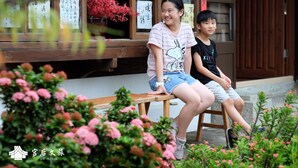 Two individuals seated on a wooden bench in front of a building with traditional Asian architectural elements.
