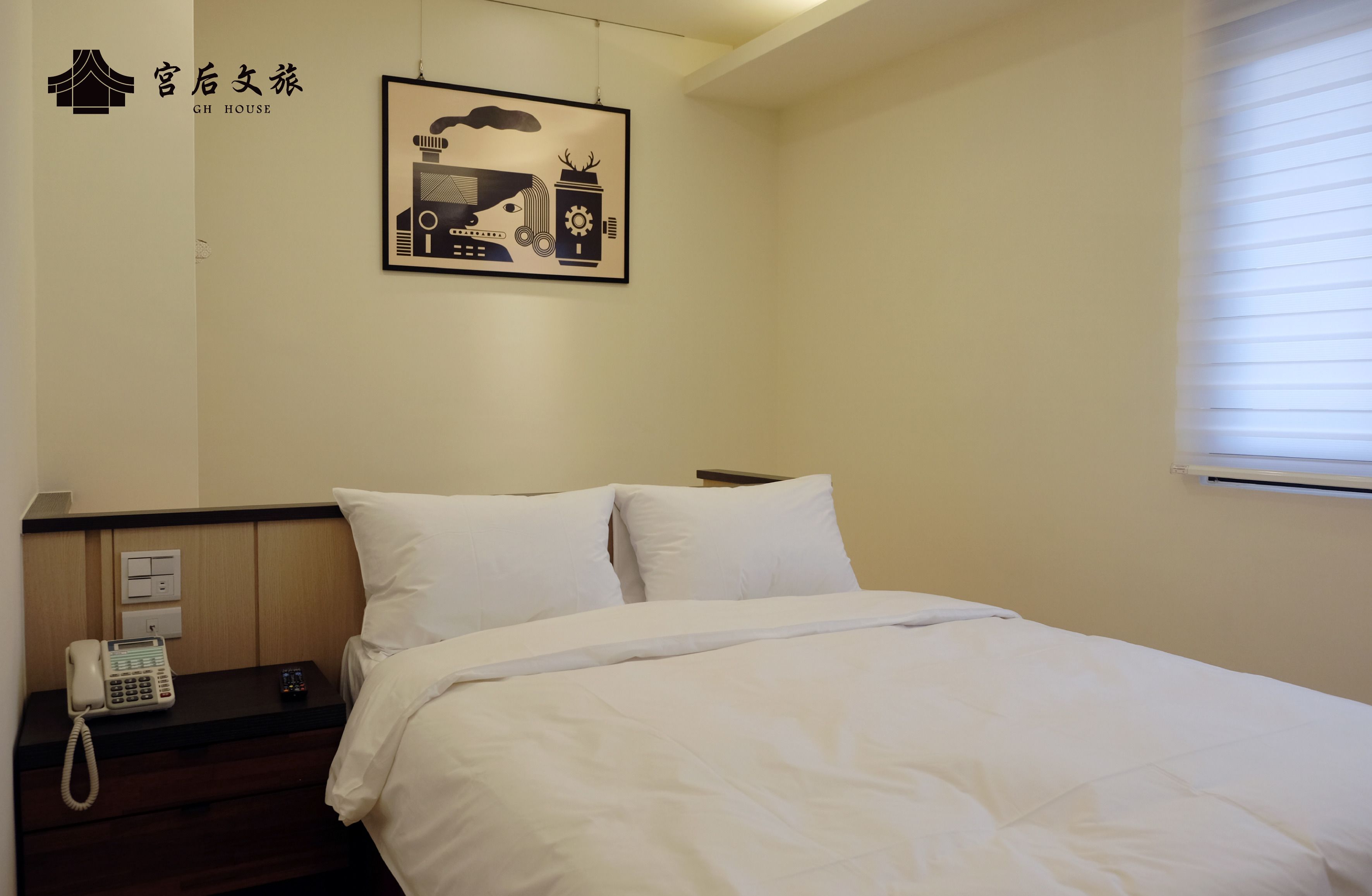 A hotel room with a bed, white bedding, a bedside phone, a framed picture on the wall, and a window with blinds.