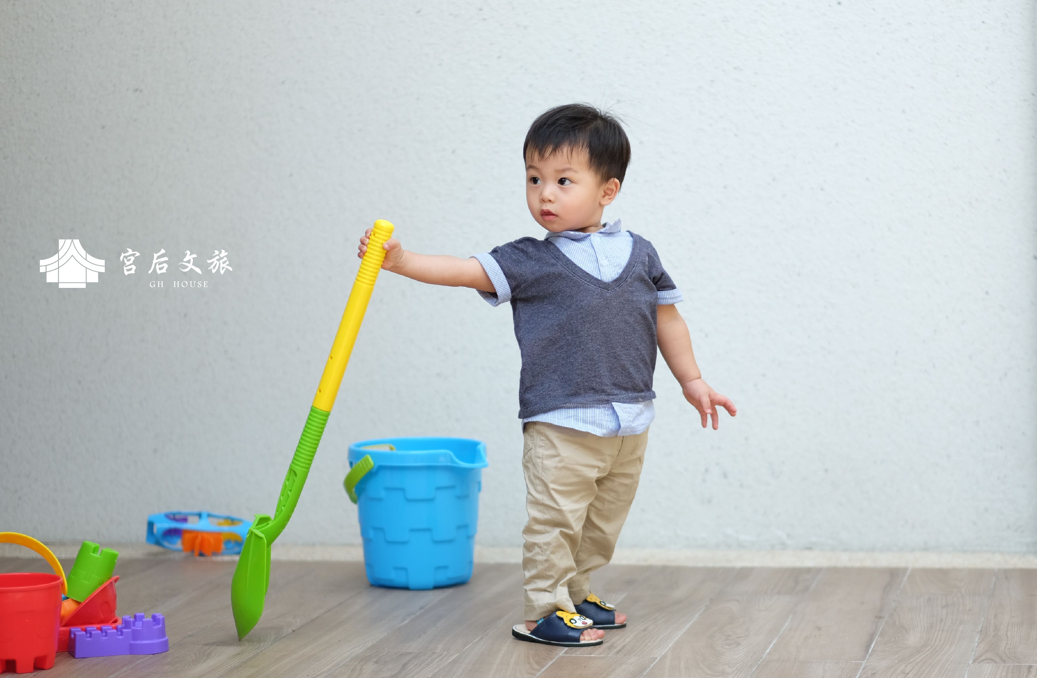 A child holding a shovel, standing next to a blue bucket and colorful toys on a wooden floor.