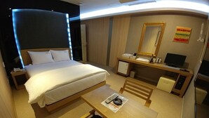 A hotel room with a bed, a desk, a chair, a small table, a mirror, a television, and a refrigerator.