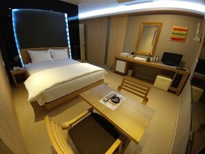 A hotel room with a bed, a desk, a chair, a small table, a mirror, a television, and a refrigerator.