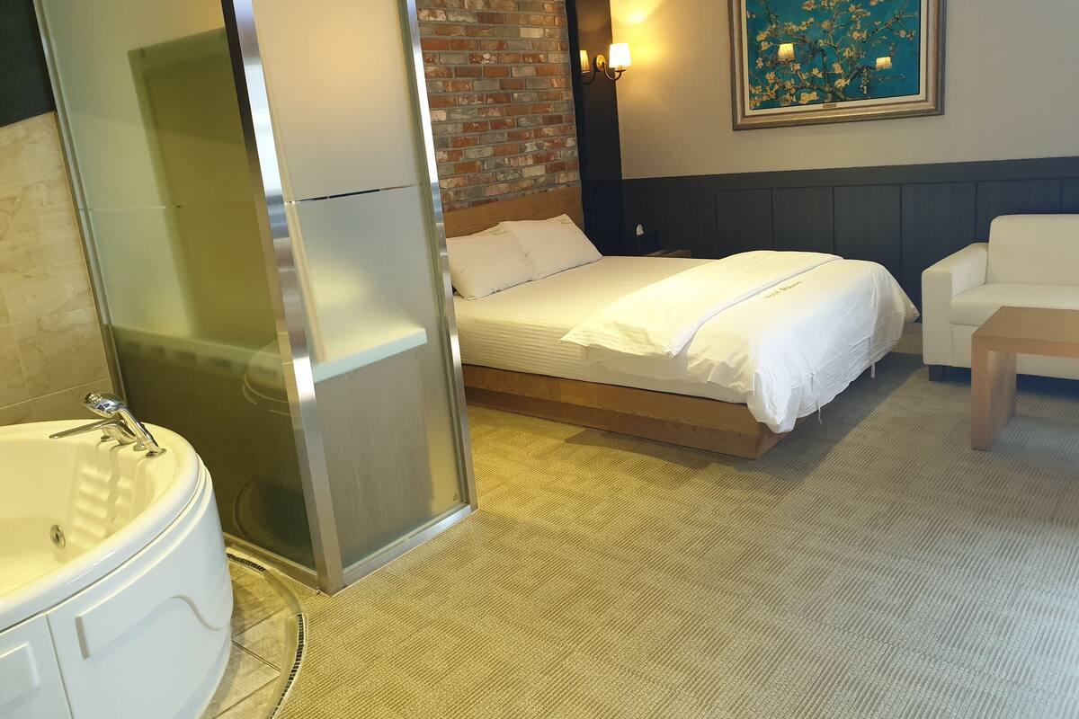 A hotel room with a bed, a white sofa, a painting on the wall, and a bathroom with a bathtub and sink.
