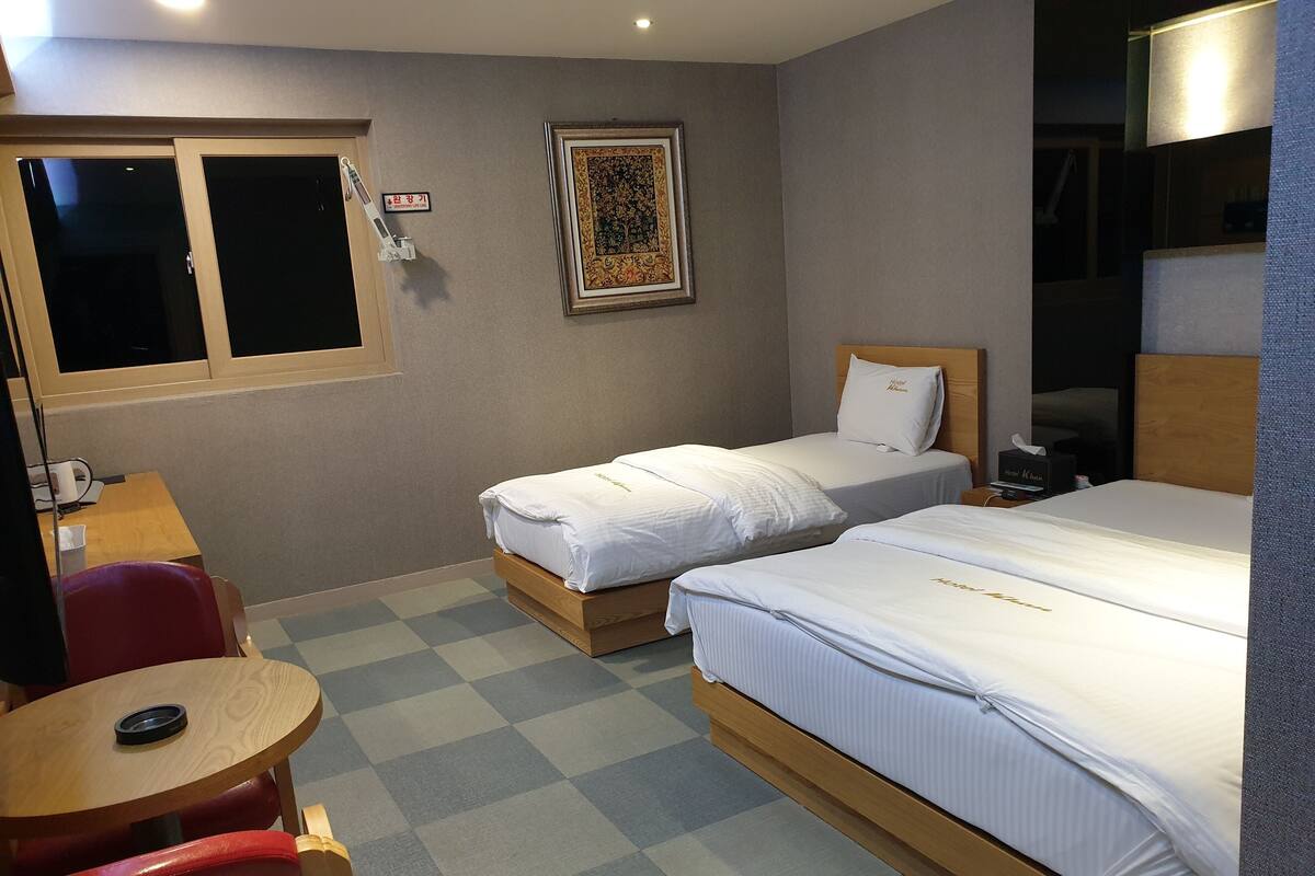 A hotel room with two single beds, a desk, a chair, a window, and a wall-mounted lamp.