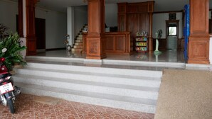 A spacious foyer with a staircase leading to a second floor, wooden columns, a motorbike parked outside, and a small indoor plant. - Dalha Renovtel Hotel (Nakhon Phanom)