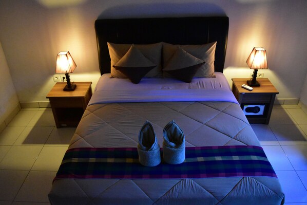 A neatly made bed with a quilted cover, two pillows, and a pair of shoes on a plaid blanket. There are two bedside tables with lamps, one of which has a remote control on it. - Meisya Cottage (Gili Trawangan)