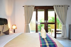 A hotel room with a bed, a bedside lamp, a window with curtains, and a view of the outdoors. - Meisya Cottage (Gili Trawangan)