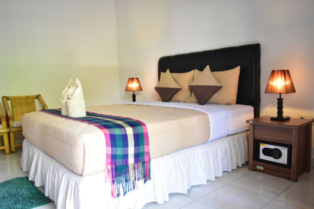 A neatly made bed with a white duvet, a colorful blanket, and decorative pillows. There are two bedside tables with lamps, a wooden chair, and a small speaker on the nightstand.