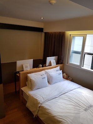 A hotel room with a bed, pillows, a nightstand, a window, and a ceiling light. - DWELL HOTEL (Busan)