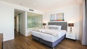 A modern bedroom with a large bed, bedside tables, a glass shower enclosure, and a painting on the wall.