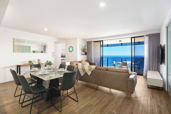 A modern living room with a dining area, a large window with a view of the sea, and a flat-screen TV mounted on the wall.