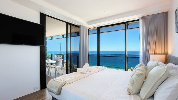 A bedroom with a large bed, a TV mounted on the wall, and a view of the sea through large windows.
