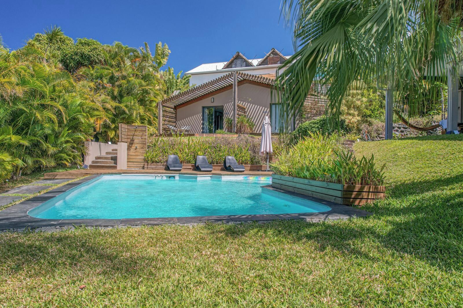 A swimming pool with lounge chairs, surrounded by lush greenery and a house with a sloped roof.