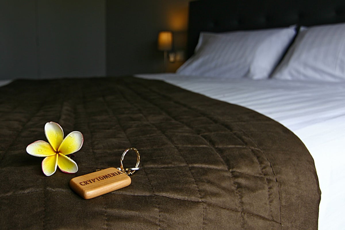 Una cama con un edredón marrón, un llavero con la etiqueta "CRYPTOMERIA" y una flor amarilla.