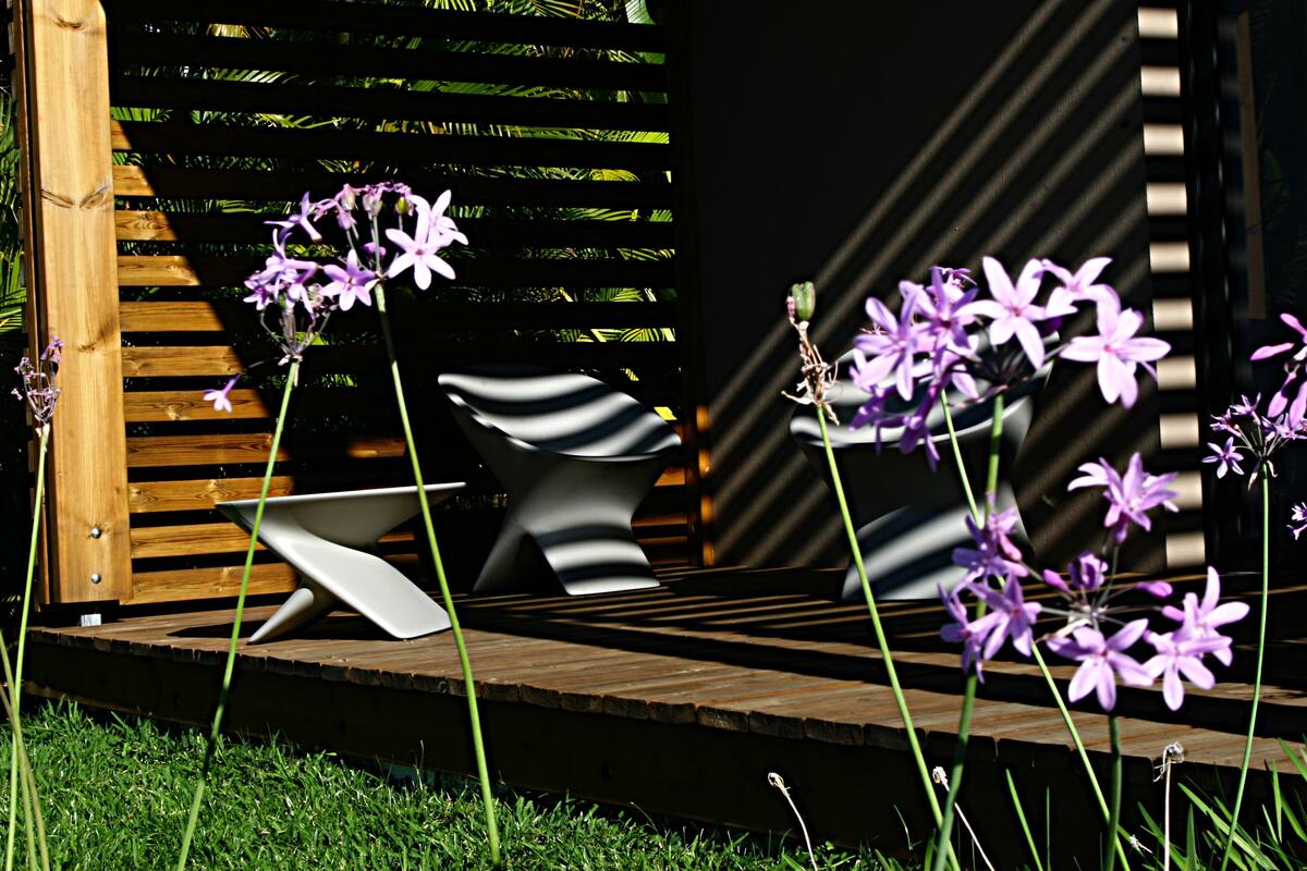 Una terraza de madera con una silla moderna y una pequeña mesa, rodeada de césped verde y flores púrpuras.