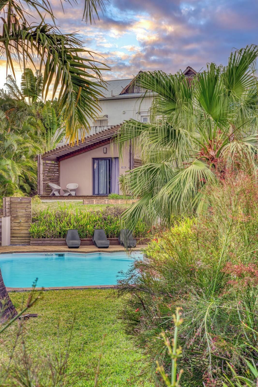 Une piscine avec des chaises longues, une maison dotée d’un patio et une végétation luxuriante.
