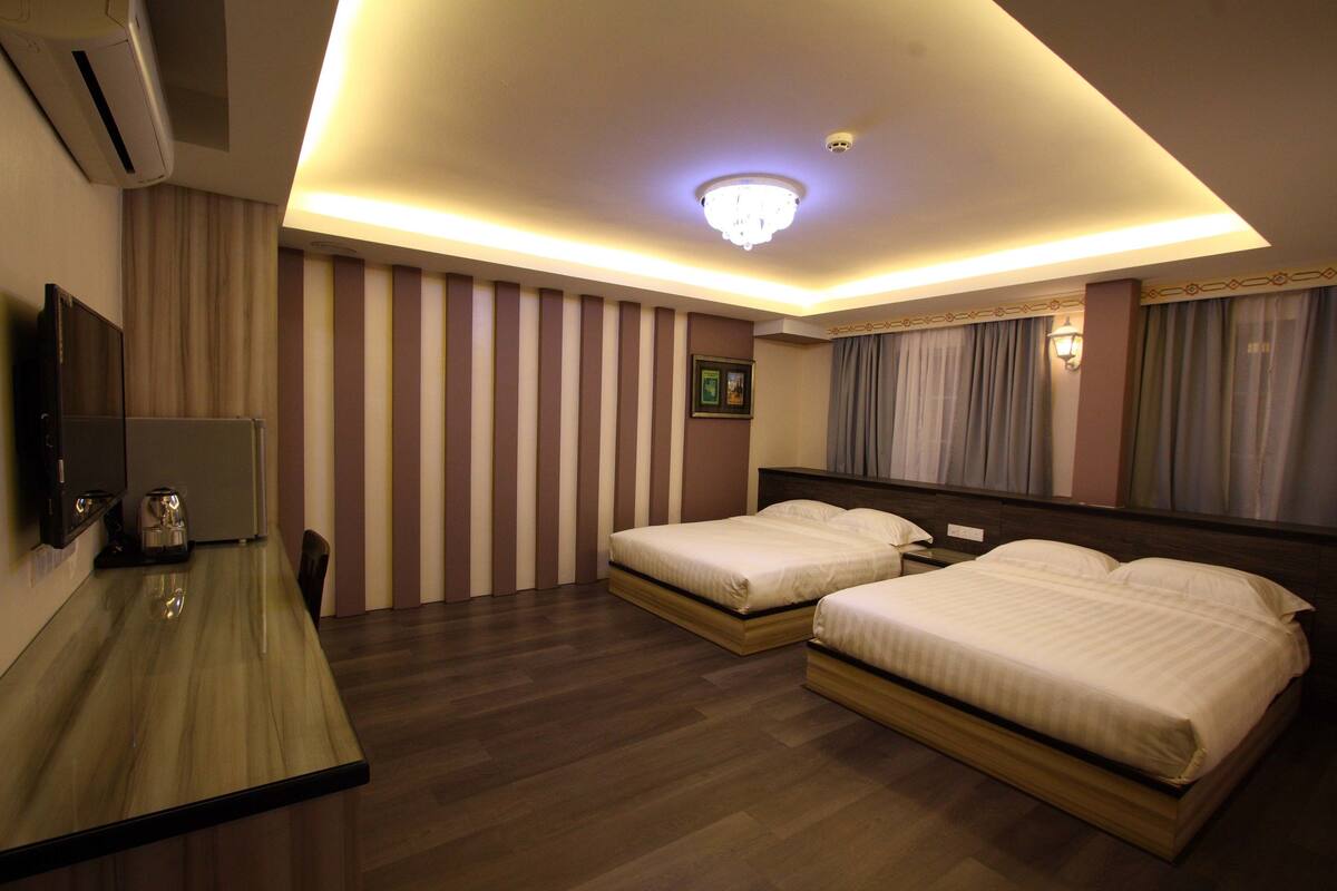 A hotel room with two beds, a desk, a TV, and a ceiling light.
