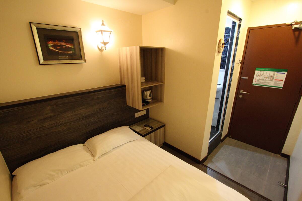 A hotel room with a bed, a nightstand, a framed picture on the wall, a shelf with a lamp, and a door with a sign.