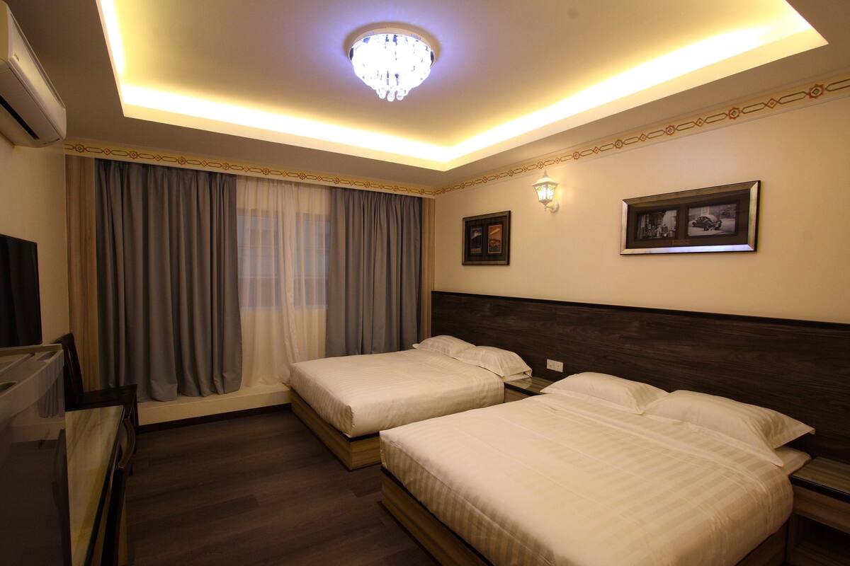 A hotel room with two beds, a ceiling light, a TV, and a window with curtains.