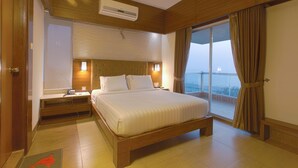 Une chambre d’hôtel dotée d’un grand lit, d’une tête de lit en bois, de tables de chevet, d’un appareil de climatisation, d’une petite table et offrant une vue sur la mer par une grande fenêtre.