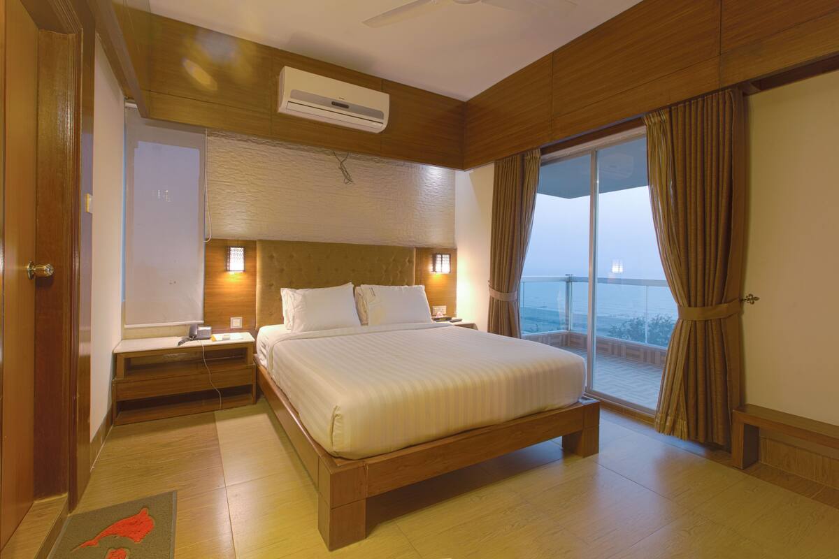 Habitación de hotel con una cama grande, cabecera de madera, mesitas de noche, un equipo de aire acondicionado y vistas al mar a través de un ventanal amplio.