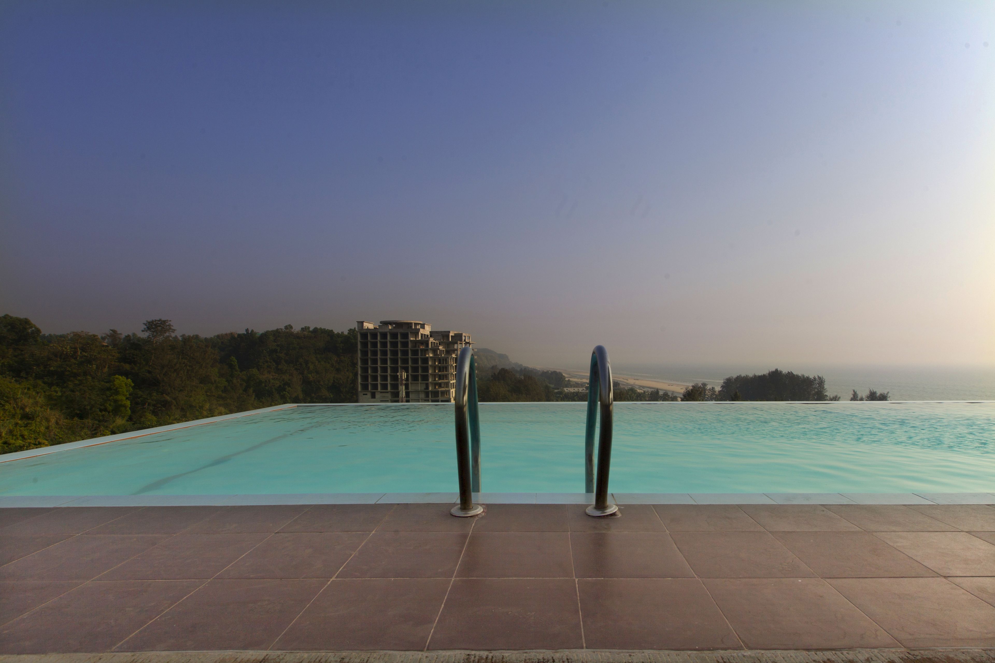 Ein Schwimmbecken mit zwei Metallhandläufen, umgeben von einer gefliesten Terrasse, mit Blick auf ein entferntes Gebäude und Bäume.