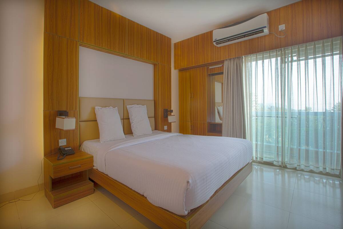 Una habitación de hotel con cabecera de madera, una cama grande con ropa blanca, una mesita de noche con lámpara y una ventana con cortinas de encaje.