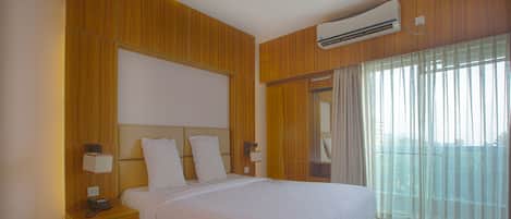 A hotel room with a wooden headboard, a large bed with white linens, a bedside table with a lamp, and a window with sheer curtains.