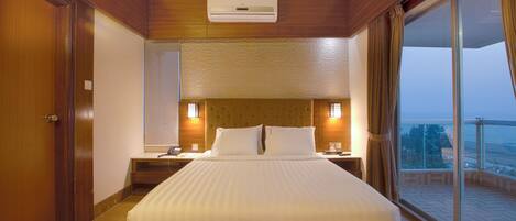 A hotel room with a large bed, wooden headboard, bedside lamps, a nightstand, and a view of the outdoors through a sliding glass door.