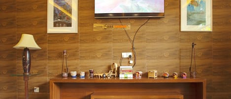 A wooden reception desk with a flat-screen TV mounted above it, displaying a program. There are framed artworks on the wall, a lamp, and a shelf with various items.