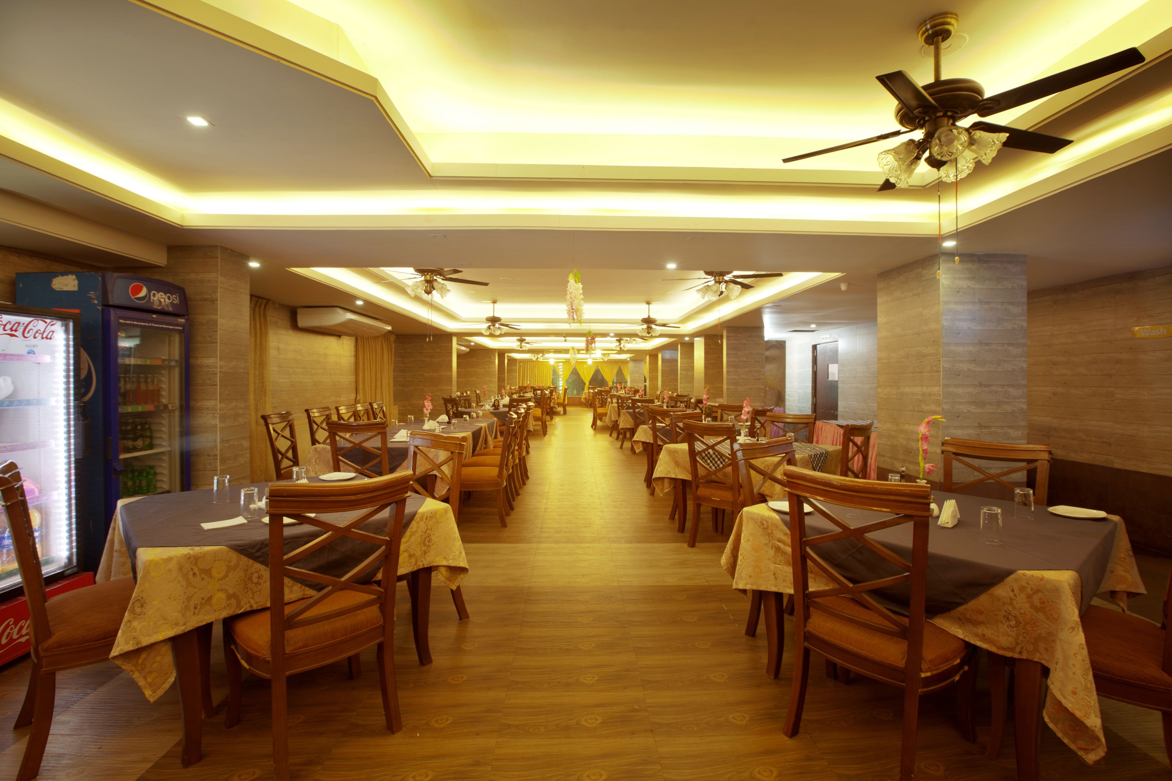 A spacious restaurant with multiple tables and chairs, ceiling fans, and a Coca-Cola refrigerator.