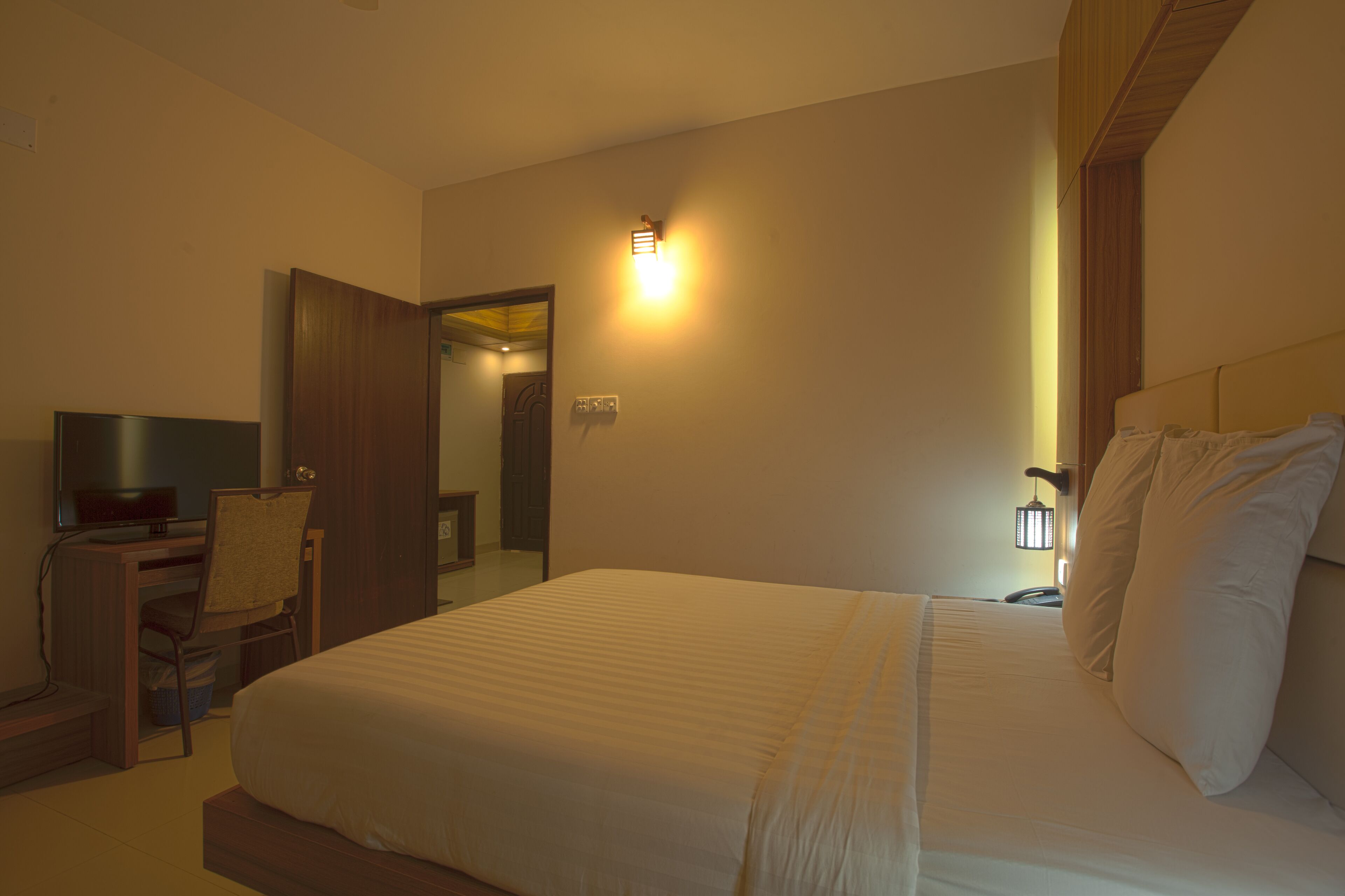 A hotel room with a bed, a desk with a chair, a television, and a wall-mounted light.