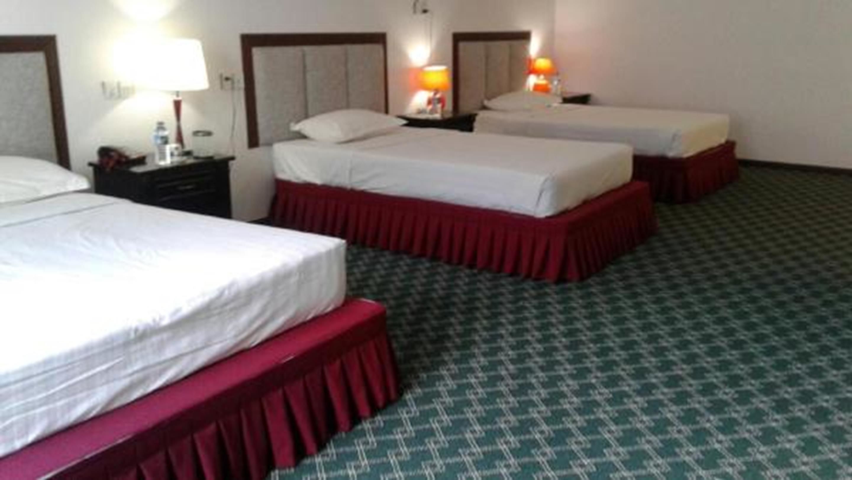 A hotel room with three beds, each with a red skirt and white linens. There are two bedside tables with lamps, and the room has a green patterned carpet.