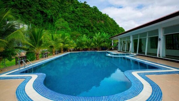 Una piscina con borde de azulejos, rodeada de exuberante vegetación y un edificio con grandes ventanales.