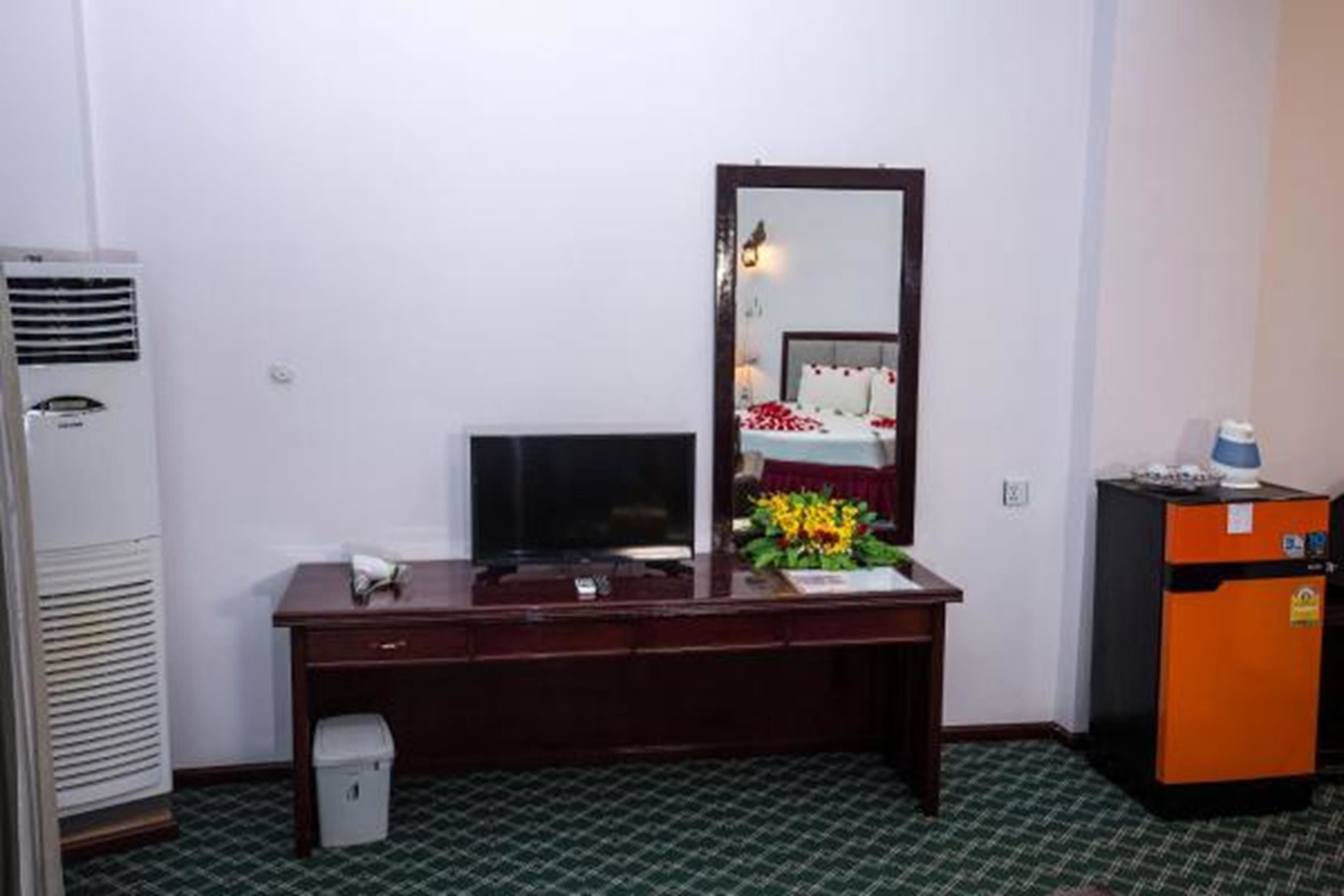 Een hotelkamer met een houten bureau, een televisie, een spiegel, een kleine koelkast en een prullenbak.
