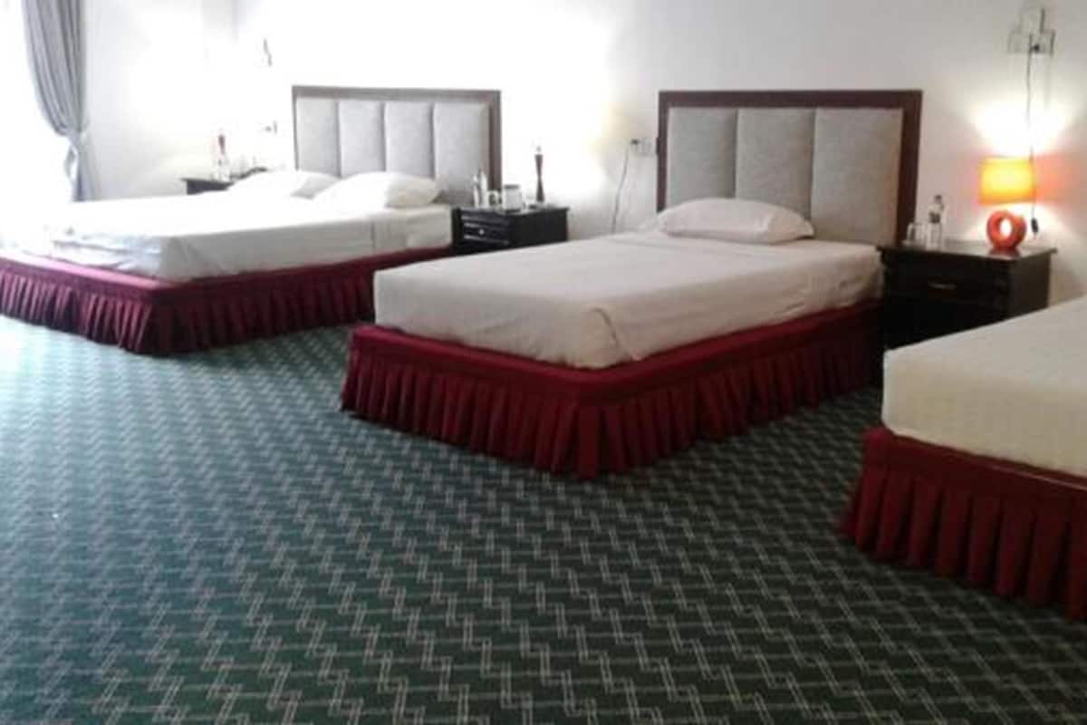 A hotel room with three beds, each with a red skirt, a headboard, and a nightstand with a lamp.