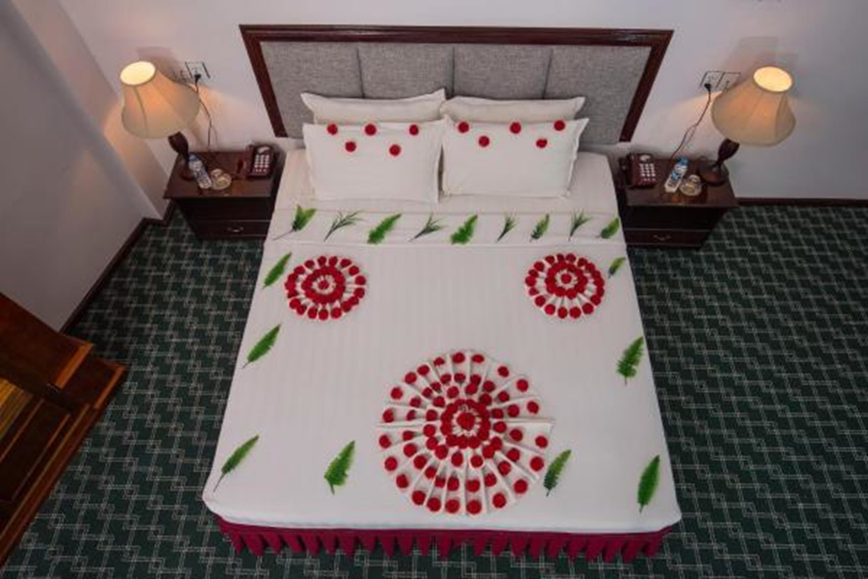 A neatly made bed with a white and red floral bedspread, two bedside tables with lamps, and a green patterned carpet.