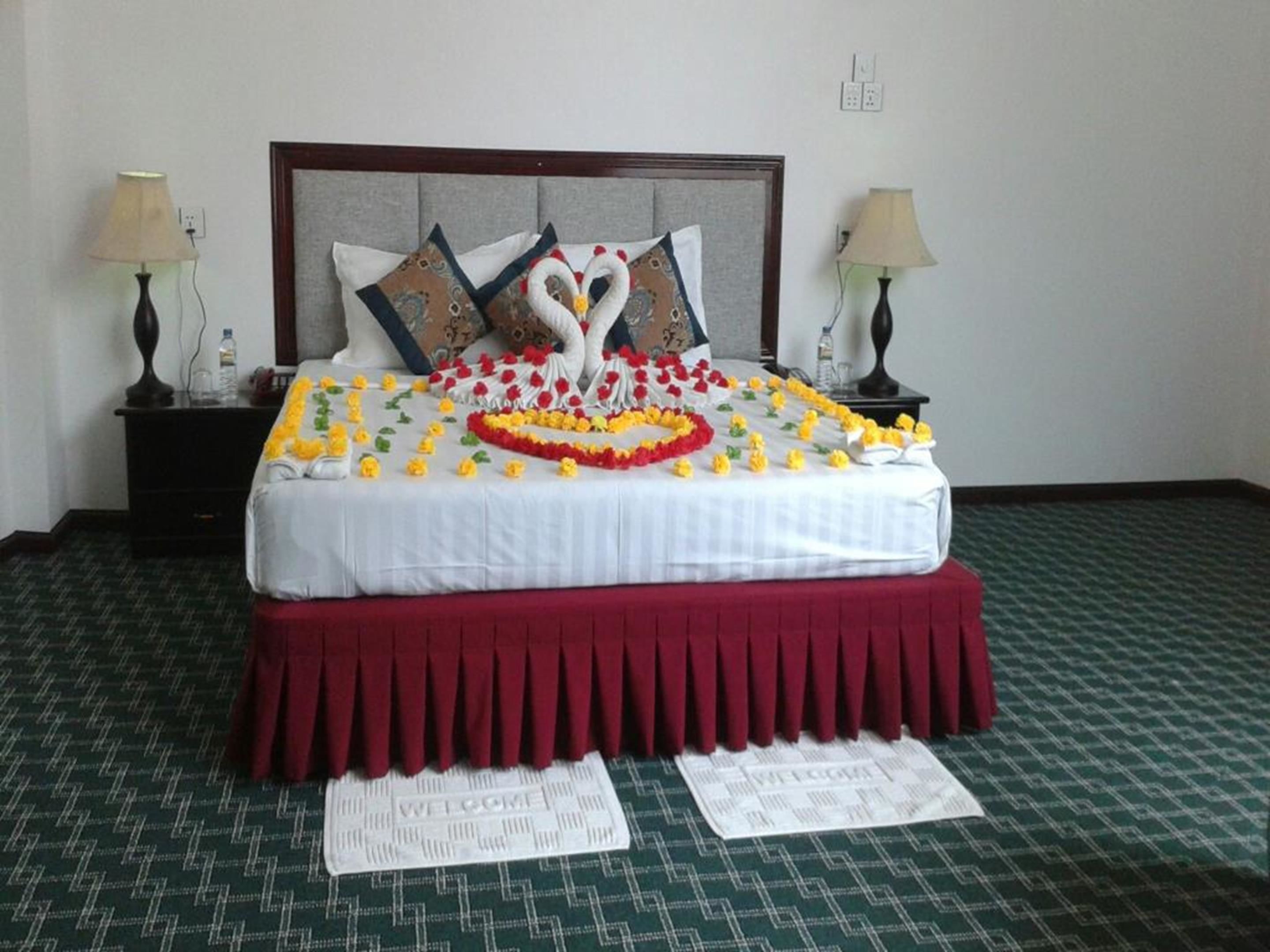 A neatly made bed with a heart-shaped decoration made of flowers and a swan design, flanked by two lamps and a nightstand with a bottle and glasses.