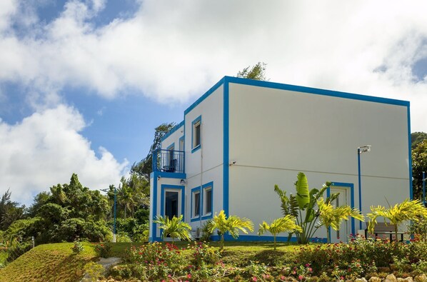 A modern two-story building with a flat roof, surrounded by lush greenery and flowering plants. - Joy House (Saipan)