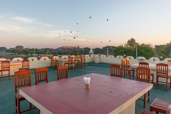 A rooftop terrace with wooden chairs and tables, overlooking a cityscape with buildings and hot air balloons in the sky.