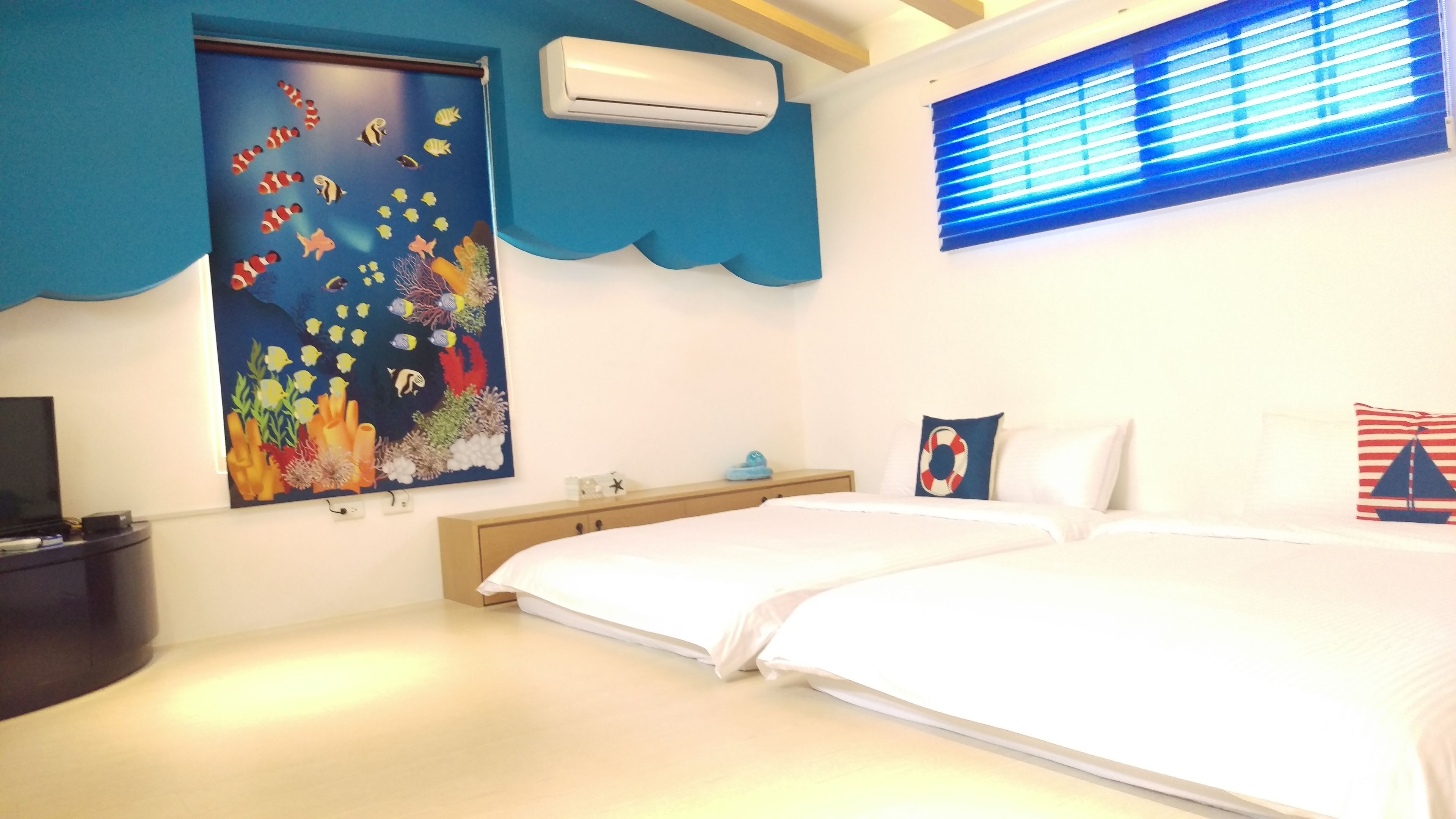 Un dormitorio con dos camas, un amplio mural en la pared con escena submarina, un equipo de aire acondicionado y una ventana con persianas.