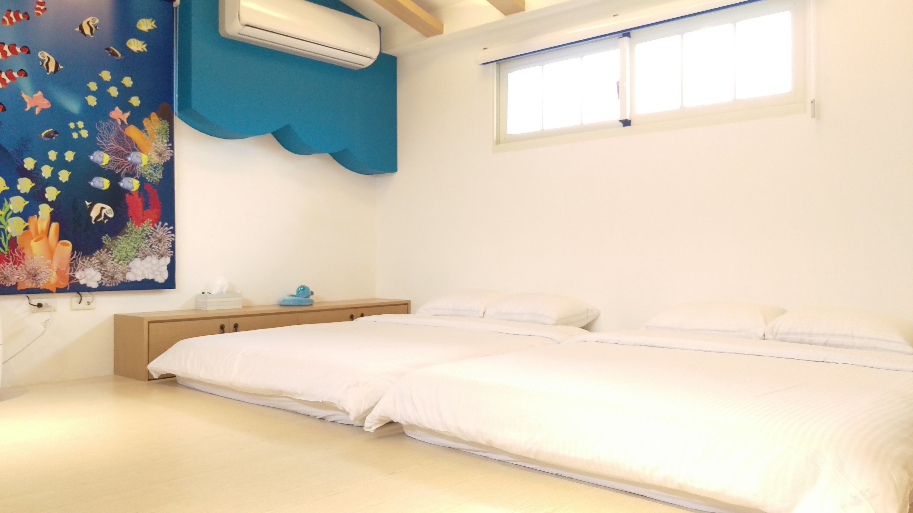 A bedroom with a bed, a nightstand, a wall-mounted air conditioner, a window, and a colorful painting of marine life.